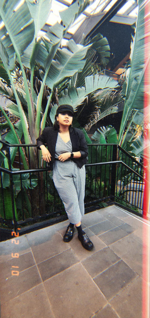 Aurelia Andrea check jumpsuit worn with black blazer, chauffer’s hat and black chunky loafers, standing on steps in front of palm trees