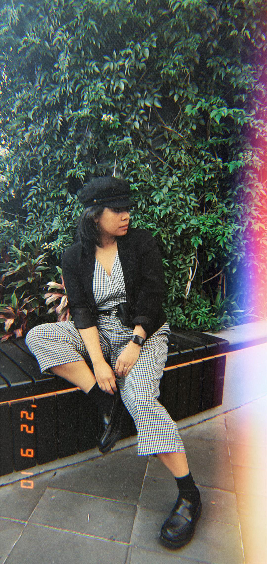 Aurelia Andrea check jumpsuit worn with black blazer, chauffer’s hat and black chunky loafers, sitting on a bench in front of green leaves